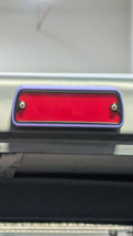 A close-up of a rectangular red taillight with a purple bumper frame and silver screws, mounted on a vehicle bumper.