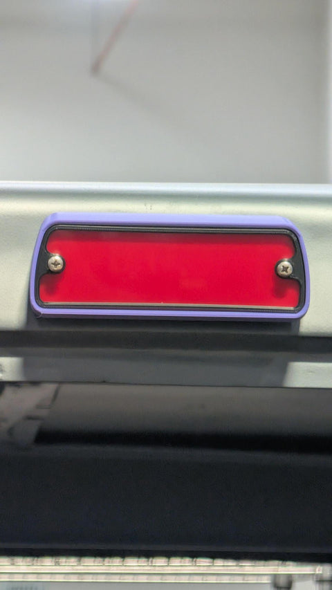 A close-up of a rectangular red taillight with a purple bumper frame and silver screws, mounted on a vehicle bumper.