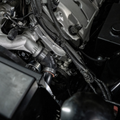 Close-up of an automotive engine bay showing a black and silver intake hose with 'Hastel' printed on it, surrounded by engine components, wiring, and hoses.