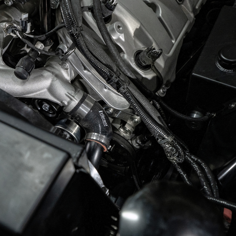 Close-up of an automotive engine bay showing a black and silver intake hose with 'Hastel' printed on it, surrounded by engine components, wiring, and hoses.