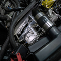 A close-up view of an automotive engine bay showing a clear plastic engine cover over a silver alternator, surrounded by black hoses, metal components, and a yellow oil cap.
