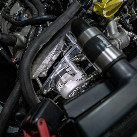 A close-up view of an automotive engine bay showing a clear plastic engine cover over a silver alternator, surrounded by black hoses, metal components, and a yellow oil cap.